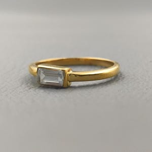 May include: A gold ring featuring a rectangular, clear gemstone. The ring's band and setting are a bright gold colour. The gemstone is set horizontally and appears faceted.