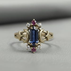 May include: A gold ring featuring a rectangular, deep blue gemstone at its center. The ring is adorned with small, round red and white gemstones, and crescent moon and star designs on the band.