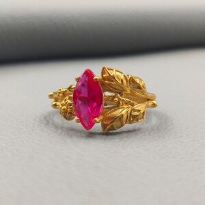 Puede incluir: Un anillo dorado con una llamativa gema rosa en forma de marquesa. El diseño del anillo incorpora intrincados patrones de hojas y flores, creando una estética inspirada en la naturaleza. La gema rosa brillante es el punto central.