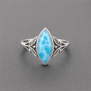 May include: A silver ring featuring a marquise-cut, light blue Larimar stone with a marbled pattern. The band is decorated with symmetrical metal leaf motifs and small spherical accents on both sides of the central gemstone setting.
