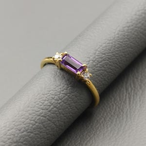 May include: A gold ring featuring a rectangular, deep purple gemstone as the centerpiece, flanked by two smaller, round, clear gemstones. The ring's band is a simple, polished gold.