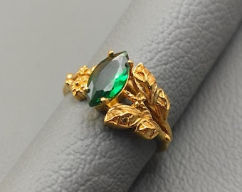 May include: A gold-colored ring featuring a marquise-cut, emerald-green gemstone. The ring has a nature-inspired design with leaf and floral details. The ring is set on a gray surface.