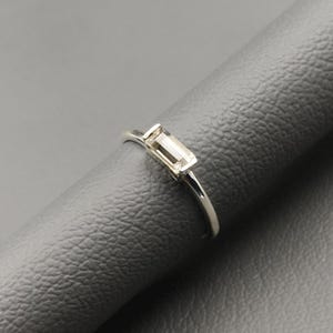 Op de afbeelding: Een zilveren ring met een rechthoekige, heldere edelsteen. De ring is geplaatst op een grijze ondergrond, wat het delicate ontwerp benadrukt. De edelsteen is het centrale punt, reflecteert licht en voegt een vleugje elegantie toe aan de sieraden.