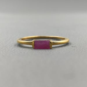 Op de afbeelding: Een goudkleurige ring met een rechthoekige, magenta gekleurde edelsteen. De ring heeft een dunne, gladde band en de edelsteen is gezet in een eenvoudige, rechthoekige bezel. De ring is waarschijnlijk gemaakt van verguld metaal.