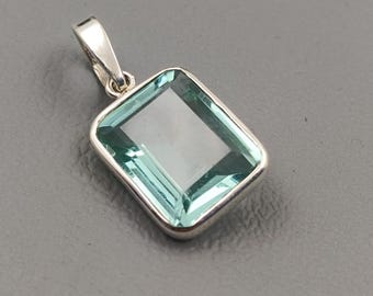 May include: A rectangular pendant with a light blue gemstone set in a silver-colored metal frame. The gemstone has a clear, transparent appearance. The pendant has a small loop at the top for a chain.