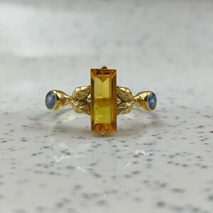 May include: A gold ring featuring a rectangular, yellow gemstone as the centerpiece, flanked by two smaller, round blue gemstones. The ring has an ornate, leaf-like design on the band.