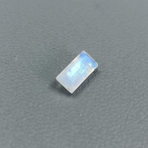 May include: A rectangular, faceted gemstone with a white body and blue iridescence. The gem is set against a gray background. The gemstone is likely a moonstone, used in jewelry.
