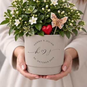 Puede incluir: Una maceta de cerámica gris claro que contiene una planta con flores blancas, un adorno de corazón rojo y una mariposa hecha con un billete de 50 euros. La maceta tiene impreso el texto "HANNA & JULIAN", "h ♡ j" y "26. OKTOBER 2026".