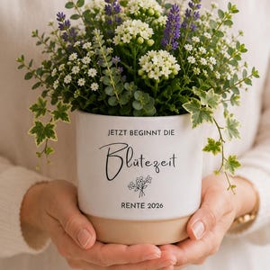 Può includere: Un vaso in ceramica bianca con base color cuoio, riempito di lavanda viola, piccoli fiori bianchi e fogliame verde. Il vaso presenta la scritta nera 'JETZT BEGINNT DIE Blütezeit RENTE 2026' e un piccolo disegno a linee di fiori.