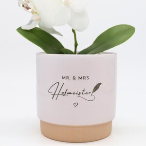 Può includere: Un'orchidea bianca in un vaso di ceramica rosa chiaro con base color sabbia. Il vaso presenta la scritta nera MR. & MRS. Hofmeister, un piccolo disegno di una piuma e un minuscolo simbolo a forma di cuore centrati sulla parte anteriore.
