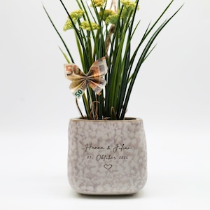 Peut inclure: Un petit pot en céramique moucheté avec des plantes vertes et des fleurs jaunes. Un papillon fait de billets de banque en euros est attaché à la plante. Le pot porte les noms "Hanna & Julian" et la date "03. Oktober 2026" avec un cœur.