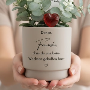 Könnte beinhalten: Ein matter beigefarbener Keramiktopf mit einer grünen Eukalyptuspflanze, kleinen weißen Blüten und einem roten Glasherz. Der Topf trägt den schwarzen Text: Danke, Franziska, dass du uns beim Wachsen geholfen hast, sowie ein kleines Herz-Symbol.