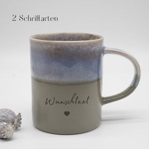 Könnte beinhalten: Eine Keramiktasse mit zweifarbiger Glasur, bestehend aus einem blaugrauen oberen Bereich und einem taupefarbenen unteren Teil. Auf der Seite ist der Text "Wunschtext" in schwarzer Schreibschrift sowie ein kleines Herz abgebildet. Oben steht "2 Schriftarten".