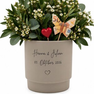 Op de afbeelding: Een matte taupekleurige keramische plantenpot met de tekst "Hanna & Julian" boven "05. Oktober 2026" en een klein hartje. De pot bevat een arrangement van groen blad, kleine witte bloemetjes, een rood hart en een vlinder gemaakt van bankbiljetten.