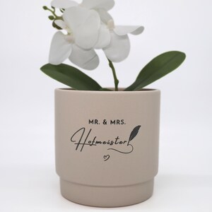 Può includere: Una pianta di orchidea bianca in un vaso cilindrico in ceramica color beige opaco. Il vaso presenta la scritta nera "MR. & MRS. Hofmeister" con una piccola icona a forma di cuore e un grafico a forma di piuma sotto il nome, su sfondo bianco.