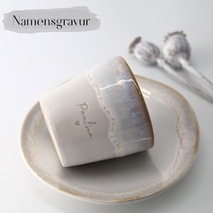 Op de afbeelding: Een keramische mok met de naam "Paulina" erop, rustend op een bijpassende schotel. De mok heeft een witte basis met iriserende randen en een lichtbruine rand. Het woord "Namensgravur" staat boven de mok.