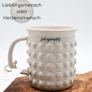 Può includere: Una tazza in ceramica beige con un motivo strutturato a forma di bolle. La tazza ha un manico e la parola "Lieblingsmensch" è stampata sul lato. Il testo "Lieblingsmensch oder Herzensmensch" è sopra la tazza.