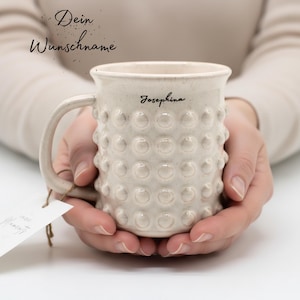 Könnte beinhalten: Eine cremefarbene Keramiktasse mit einer strukturierten Oberfläche aus erhabenen Punkten. Die Tasse hat einen Henkel und den Namen Josephina in schwarzer Schreibschrift am Rand. Der Text Dein Wunschname steht darüber in schwarzer Schrift.