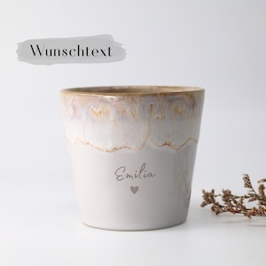 Peut inclure: Un mug en céramique avec une base lisse de couleur claire et un bord émaillé réactif dans des tons beige et blanc. L'avant du mug présente le prénom Emilia dans une police cursive au-dessus d'un petit cœur. Le texte Wunschtext est visible.