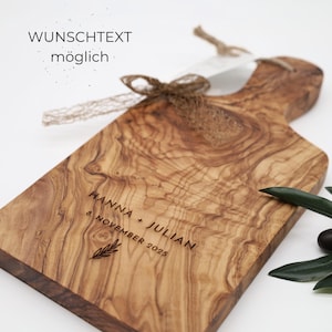 May include: A rectangular olive wood serving board with a handle, featuring a natural wood grain pattern. The board is engraved with the text HANNA + JULIAN, 8. NOVEMBER 2025, and a small leaf icon. A brown jute ribbon is tied around the handle.
