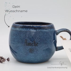 Könnte beinhalten: Eine runde, dunkelblaue Keramiktasse mit gesprenkelter Glasur und dunklerem Rand. Der Name Amélie ist in Schwarz auf der Vorderseite aufgedruckt, ergänzt durch ein kleines Blumensymbol. Der Text Dein Wunschname und Geschenkverpackung ist zu sehen.