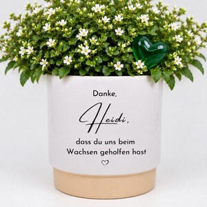 Può includere: Un vaso per piante in ceramica bianca con base color sabbia, contenente una pianta verde con piccoli fiori bianchi e un cuore in vetro verde. Il vaso riporta il testo nero: Danke, Heidi, dass du uns beim Wachsen geholfen hast, con un piccolo cuore.