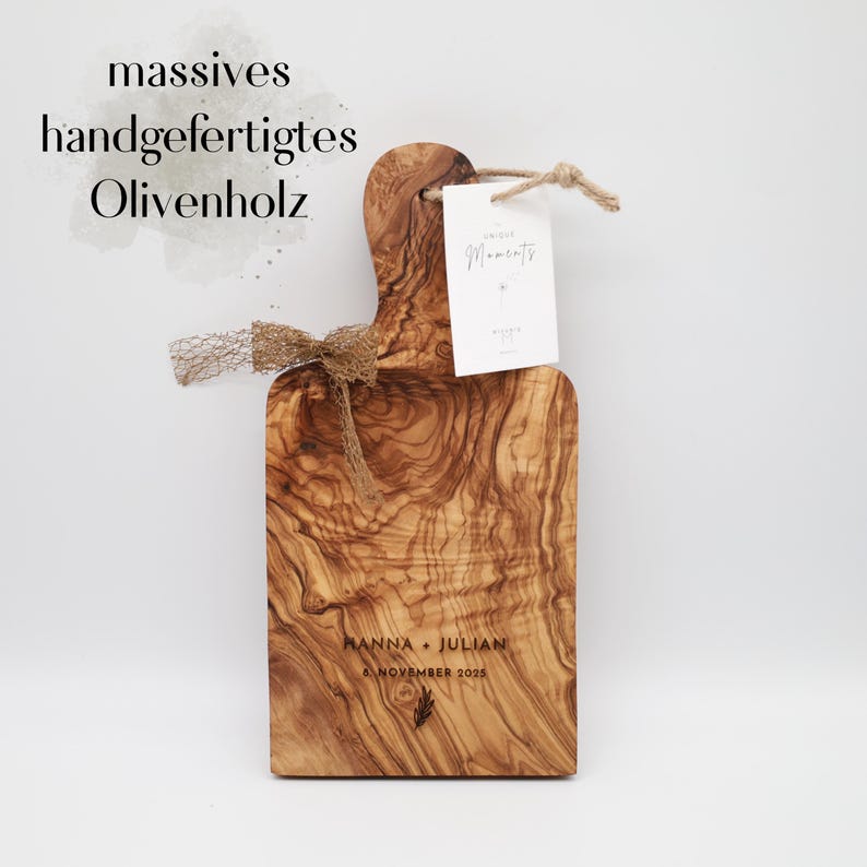 May include: A rectangular olive wood serving board with a handle, featuring a natural wood grain pattern. The board is engraved with the text HANNA + JULIAN and 8 NOVEMBER 2025. It is decorated with a brown mesh ribbon and a white tag labeled Unique Moments.