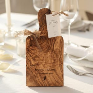 May include: A rectangular wooden serving board with a handle, featuring a natural wood grain pattern. The board is engraved with the text HANNA + JULIAN and 8 NOVEMBER 2025. A white tag tied with twine reads UNIQUE MOMENTS, and a burlap bow is attached.