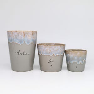 May include: Three ceramic cups of varying sizes with a smooth gray base and a textured, dripped glaze rim in shades of blue, white, and tan. The largest cup features the name Christian, while the middle cup displays the name Lea with a small heart.