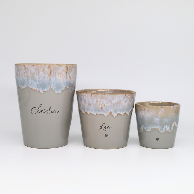 May include: Three ceramic cups of varying sizes with a smooth gray base and a textured, dripped glaze rim in shades of blue, white, and tan. The largest cup features the name Christian, while the middle cup displays the name Lea with a small heart.