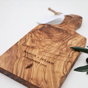 May include: A rectangular olive wood serving board with a handle, featuring a natural wood grain pattern. The board is engraved with the text HANNA + JULIAN, the date 8. NOVEMBER 2025, and a small leaf icon. A jute string is attached to the handle.