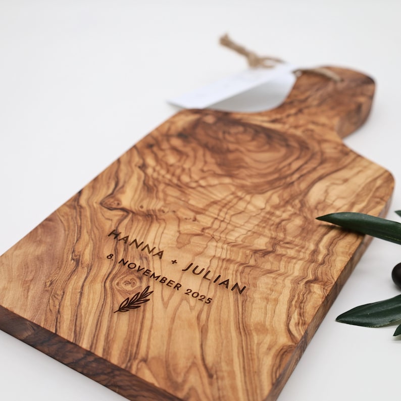 May include: A rectangular olive wood serving board with a handle, featuring a natural wood grain pattern. The board is engraved with the text HANNA + JULIAN, the date 8. NOVEMBER 2025, and a small leaf icon. A jute string is attached to the handle.