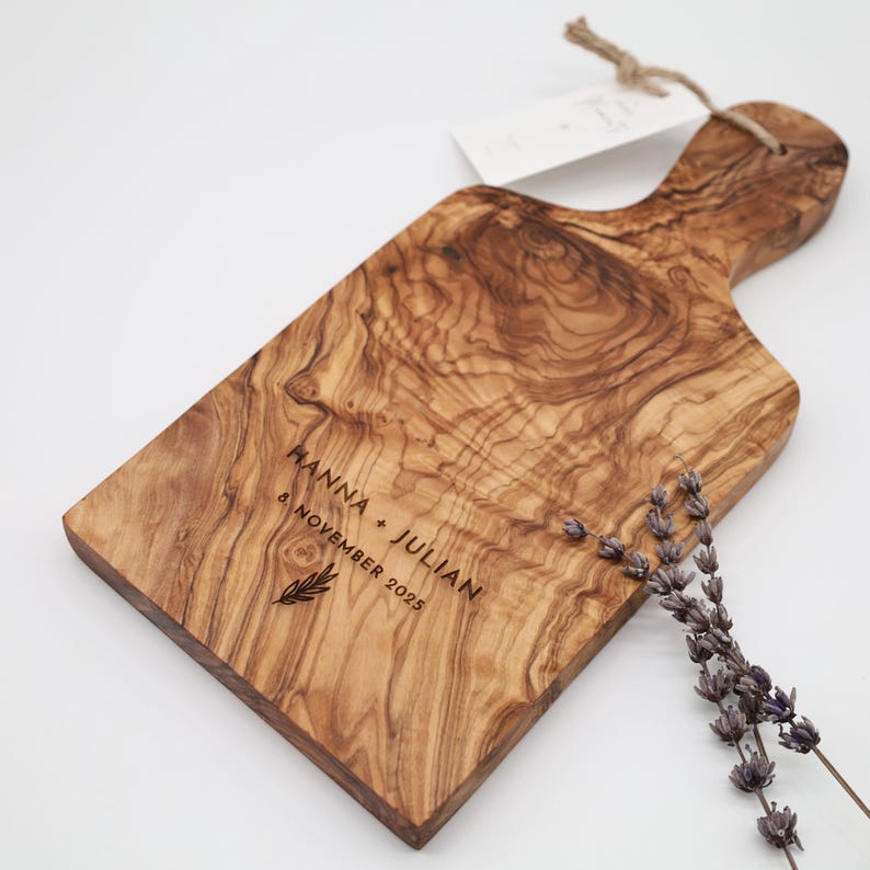 May include: A rectangular olive wood serving board with a handle, featuring a natural wood grain pattern. The board is engraved with the text "HANNA + JULIAN" and "8. NOVEMBER 2025" above a small leaf icon. A sprig of dried lavender rests on the board.