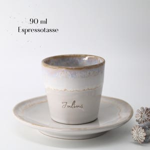 May include: A ceramic espresso cup and saucer set. The cup is white with a light brown rim and a gray base, and the saucer is white. The cup has the name "Julius" printed on it. The text "90 ml Espressotasse" is above the cup.