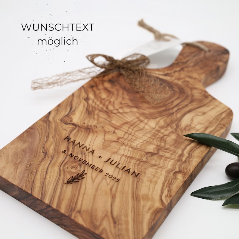 May include: A rectangular olive wood serving board with a handle, featuring a natural wood grain pattern. The board is engraved with the text HANNA + JULIAN, 8. NOVEMBER 2025, and a small leaf icon. A brown jute ribbon is tied around the handle.