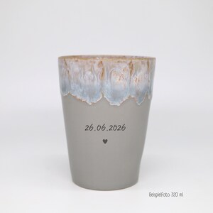 May include: A ceramic tumbler with a matte gray base and a reactive glaze rim in shades of blue, white, and brown. The front features the printed date 26.06.2026 above a small heart icon. The text "Beispielfoto 320 ml" is visible in the bottom corner.