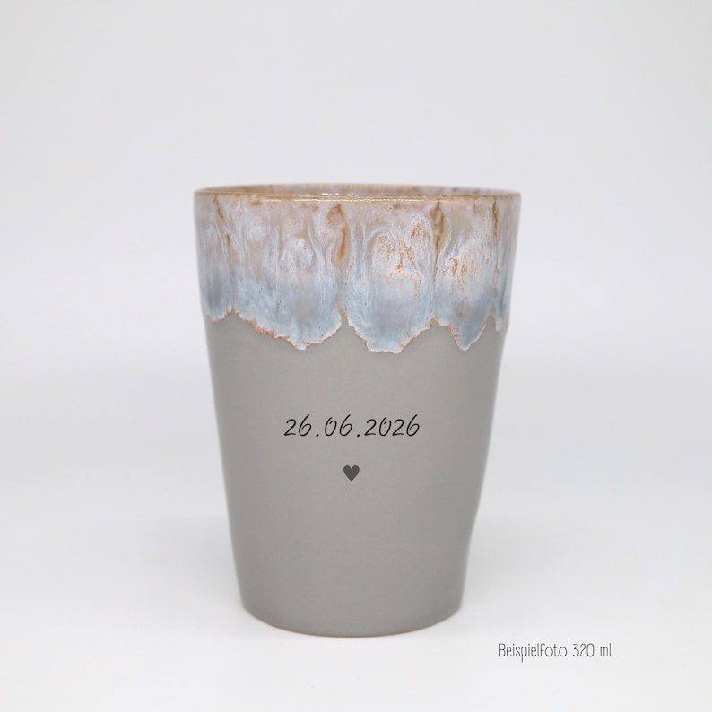 May include: A ceramic tumbler with a matte gray base and a reactive glaze rim in shades of blue, white, and brown. The front features the printed date 26.06.2026 above a small heart icon. The text "Beispielfoto 320 ml" is visible in the bottom corner.