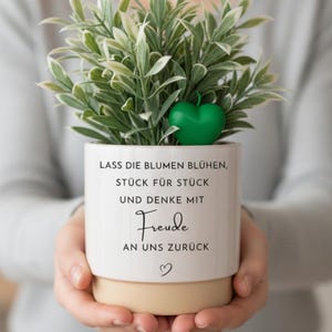 Könnte beinhalten: Ein weißer Keramikübertopf mit beigem Boden, der eine grüne Pflanze und ein kleines grünes herzförmiges Ornament enthält. Der Topf trägt den deutschen Text: LASS DIE BLUMEN BLÜHEN, STÜCK FÜR STÜCK UND DENKE MIT Freude AN UNS ZURÜCK, sowie ein kleines Herzsymbol.