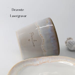 May include: A ceramic mug with a light gray base and a gradient rim of blue and brown. The name "Paulina" is written on the side in a delicate script, with a small heart below. The text "Dezente Lasergravur" is above the mug.