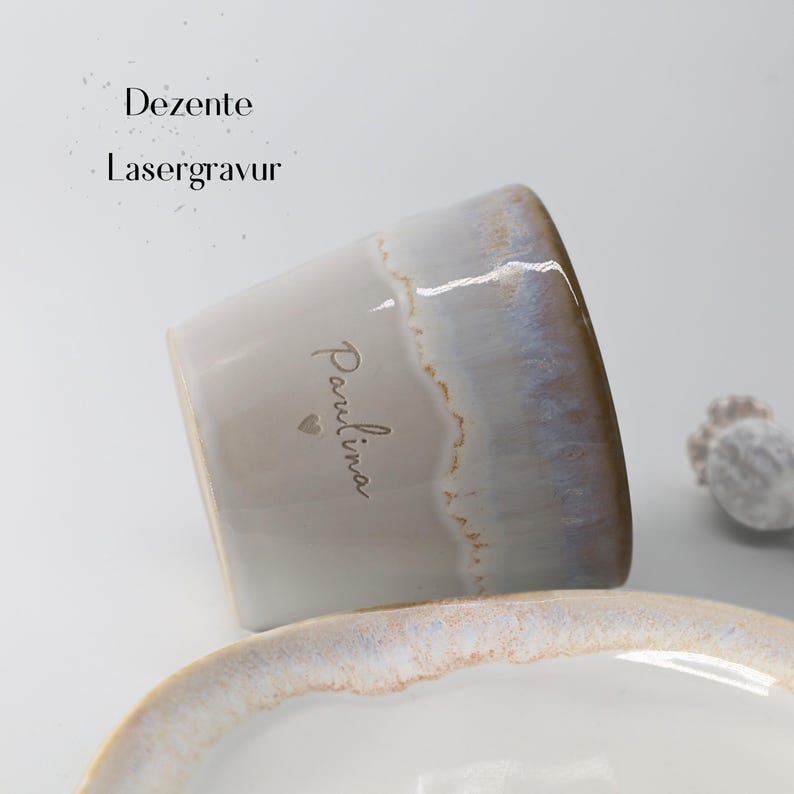 May include: A ceramic mug with a light gray base and a gradient rim of blue and brown. The name "Paulina" is written on the side in a delicate script, with a small heart below. The text "Dezente Lasergravur" is above the mug.