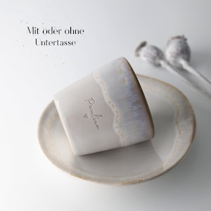 May include: A ceramic mug with a matching saucer. The mug is off-white with a glazed rim and the name "Paulina" is written on the side. The text "Mit oder ohne Untertasse" is above the mug. Dried seed pods are in the background.
