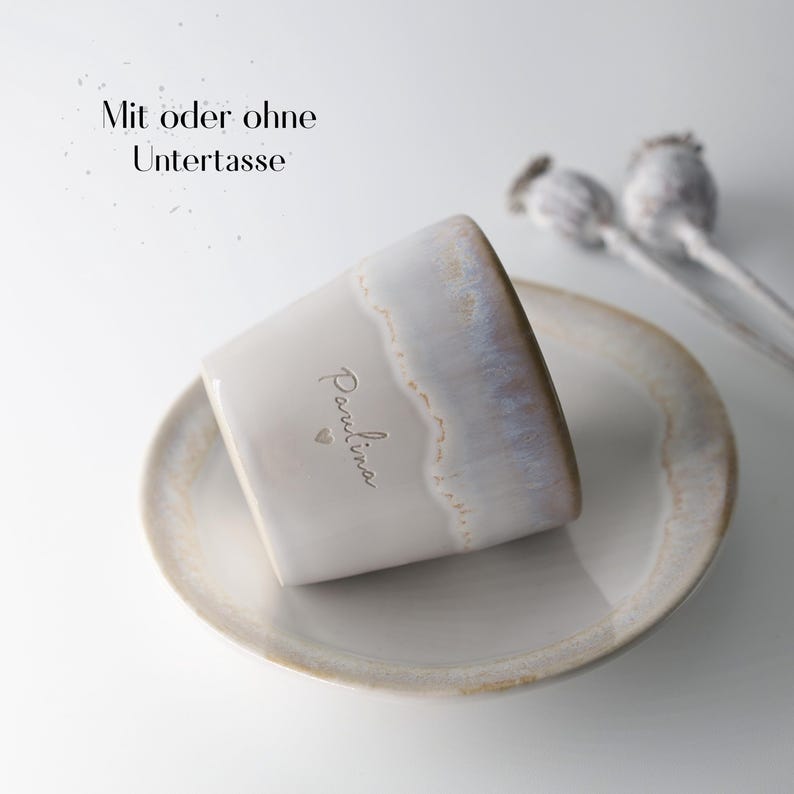 May include: A ceramic mug with a matching saucer. The mug is off-white with a glazed rim and the name "Paulina" is written on the side. The text "Mit oder ohne Untertasse" is above the mug. Dried seed pods are in the background.