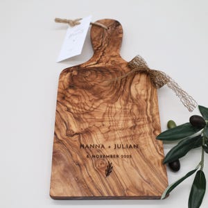 May include: A rectangular olive wood serving board with a handle, featuring a natural wood grain pattern. The board is engraved with the text HANNA + JULIAN, 8. NOVEMBER 2025, and a small leaf icon. It includes a jute tag and a gold mesh ribbon.