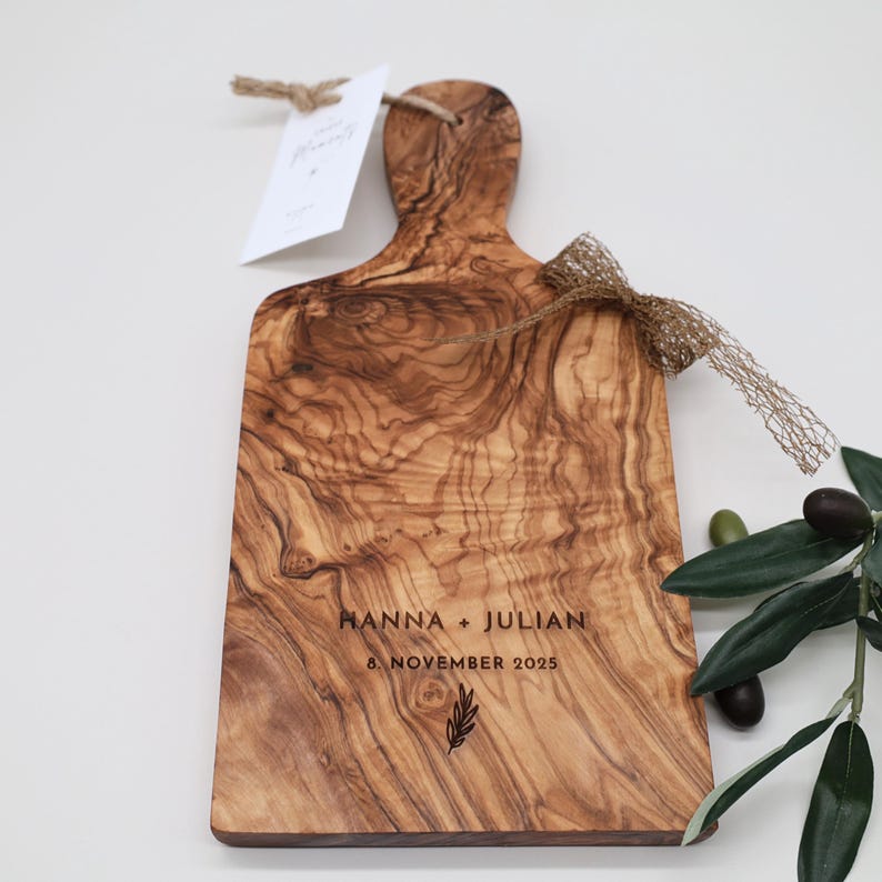 May include: A rectangular olive wood serving board with a handle, featuring a natural wood grain pattern. The board is engraved with the text HANNA + JULIAN, 8. NOVEMBER 2025, and a small leaf icon. It includes a jute tag and a gold mesh ribbon.