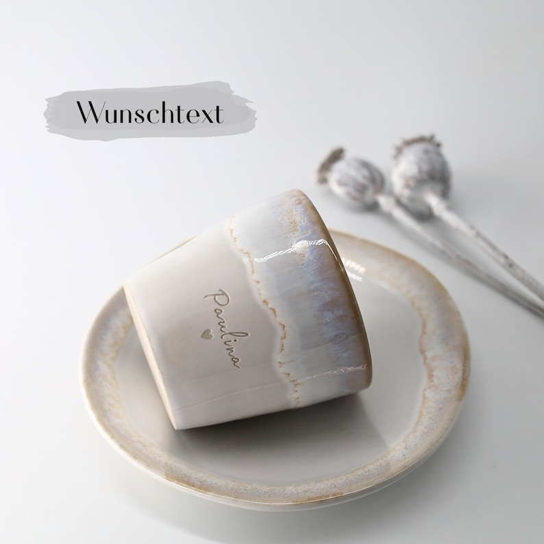 May include: A ceramic mug with a personalized name, "Paulina", and a heart symbol, resting on a matching saucer. The mug has a pearlescent glaze with a light brown rim. The text "Wunschtext" is above the mug.