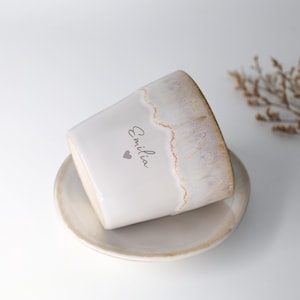 May include: A white ceramic planter with a matching saucer. The planter has a textured, off-white finish with a light brown rim. The name "Emilia" and a heart are printed on the side. A decorative item for the home.