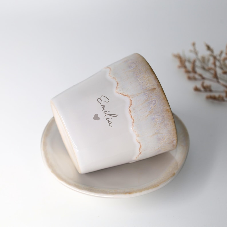 May include: A white ceramic planter with a matching saucer. The planter has a textured, off-white finish with a light brown rim. The name "Emilia" and a heart are printed on the side. A decorative item for the home.