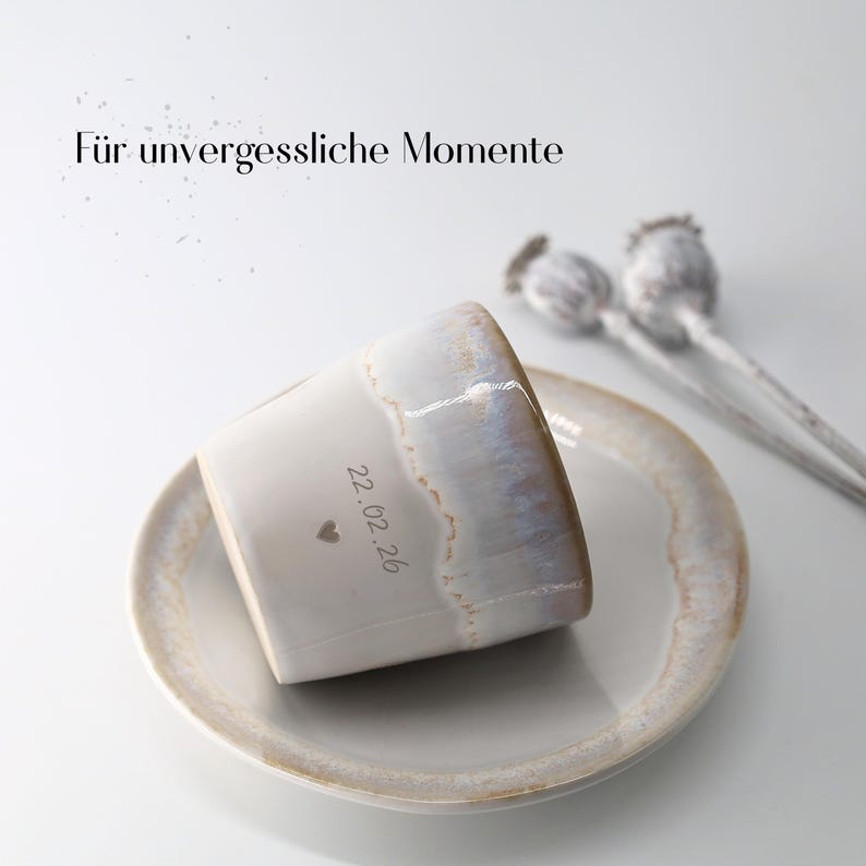 May include: A white ceramic mug with a light brown rim and a matching saucer. The mug has the date "22.02.26" and a heart symbol etched on its side. The text "F&uuml;r unvergessliche Momente" is at the top.