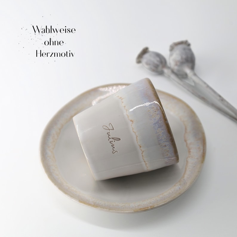 May include: A white ceramic cup with a light blue and brown rim, resting on a matching saucer. The cup has the name "Julius" written on it. The image also includes two dried seed pods and text that reads "Wahlweise ohne Herzmotiv."
