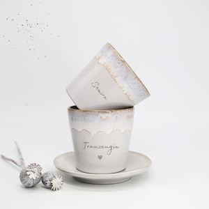 May include: Two white ceramic cups with a light brown rim and the names "Samira" and "Trauzeugin" printed on them. One cup is stacked on top of the other. A small saucer is under the bottom cup. A decorative element is on the left.
